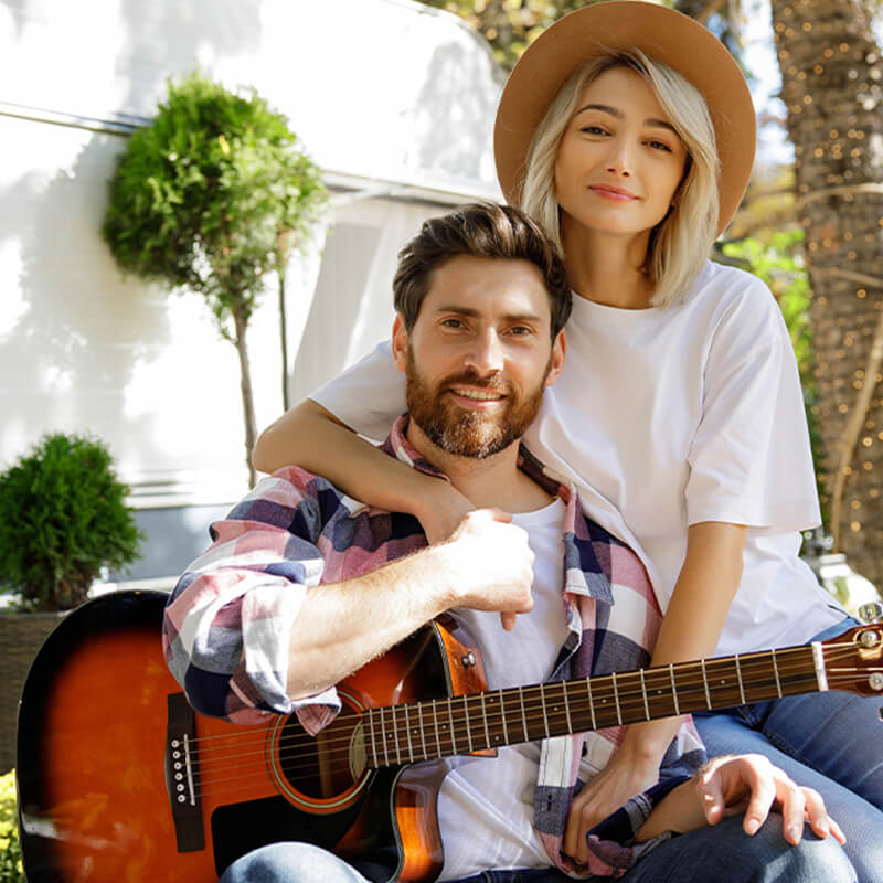 A male, and female couple setting together.  Male is holding a guitar.  Female is wearing a big hat